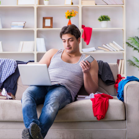 Unkempt man on a Couple online speed date. He is sitting in a messy room with a computer in his lap and a phone in his hand.