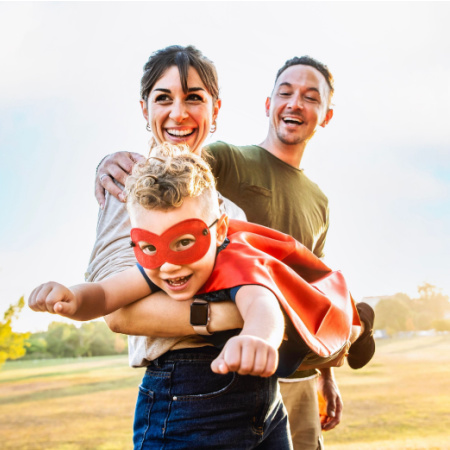 Single mom holding her kid (who's dressed as a superhero) while her new partner looks on smiling with his arm around her.