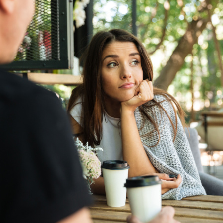 Unhappy woman on a bad first date sits at a table with a coffee cup. Only her date's shoulder is visible. 