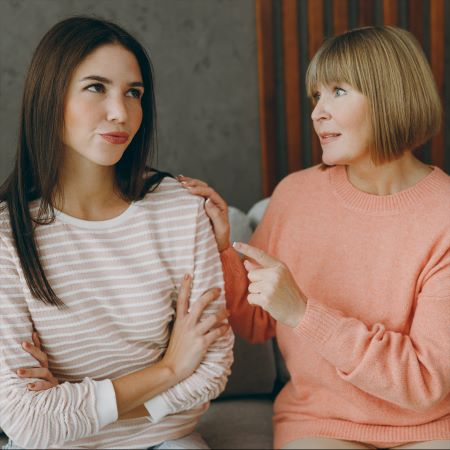 Mother and adult daughter sitting together. Mother is asking prying questions about daughter's love life while daughter sits with annoyed look on her face.