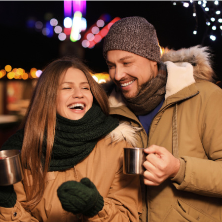 Man and woman on a holiday-themed date. They are bundled up for winter and holding mugs of cocoa. Holiday lights twinkle in the background.