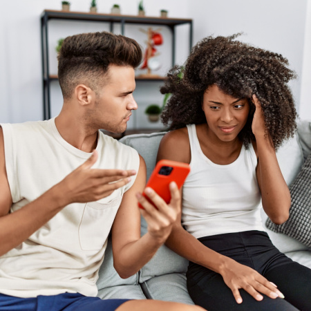Man and woman sitting on couch. Man appears distressed as he looks at his partner's phone, discovering she's cheating. The woman looks guilty and sad.