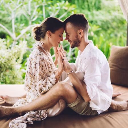 Mindful man and woman sit straddling one another, foreheads and palms pressed together, with their eyes closed.